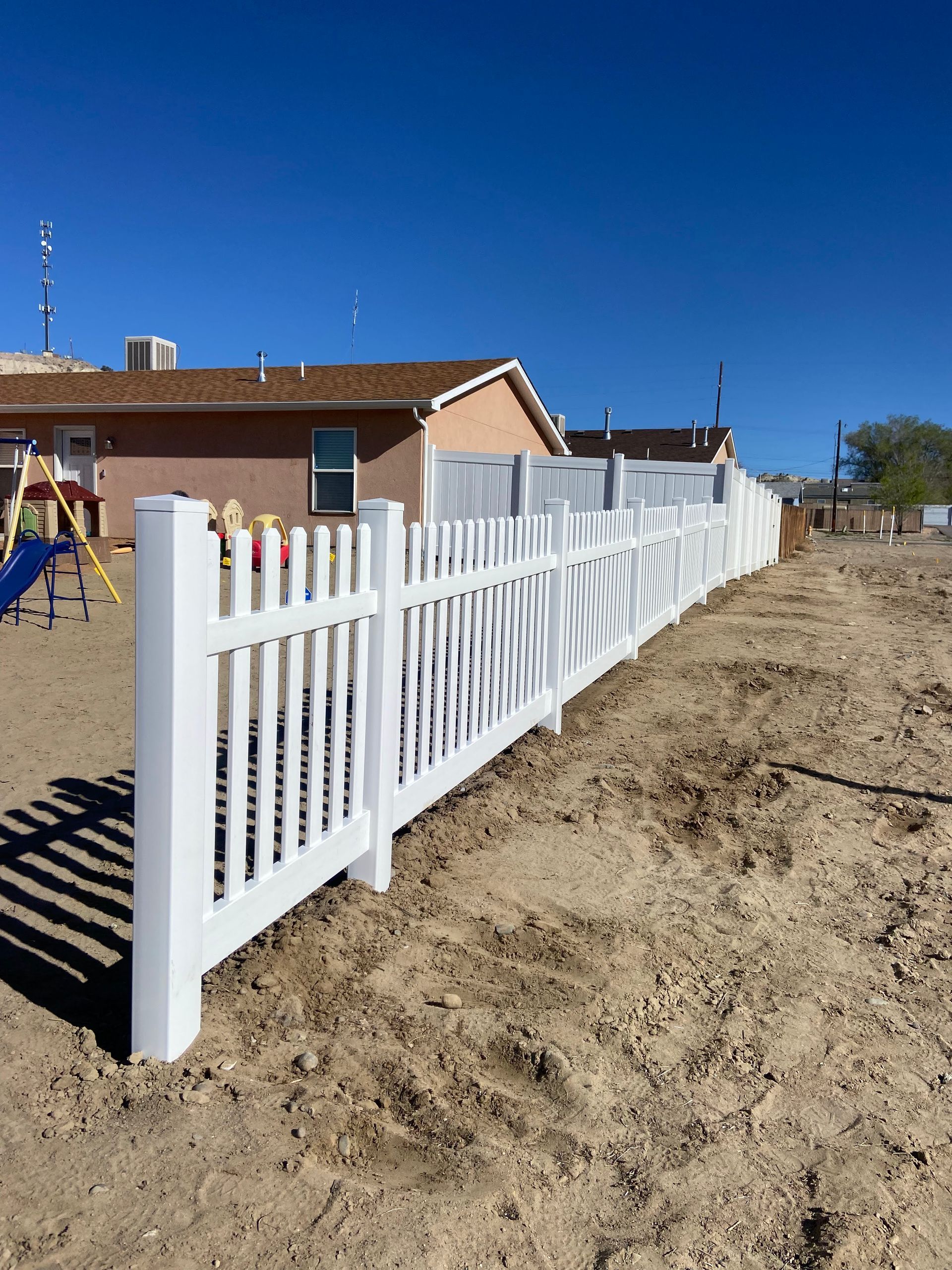 A white picket fence is sitting on top of a dirt field next to a playground.