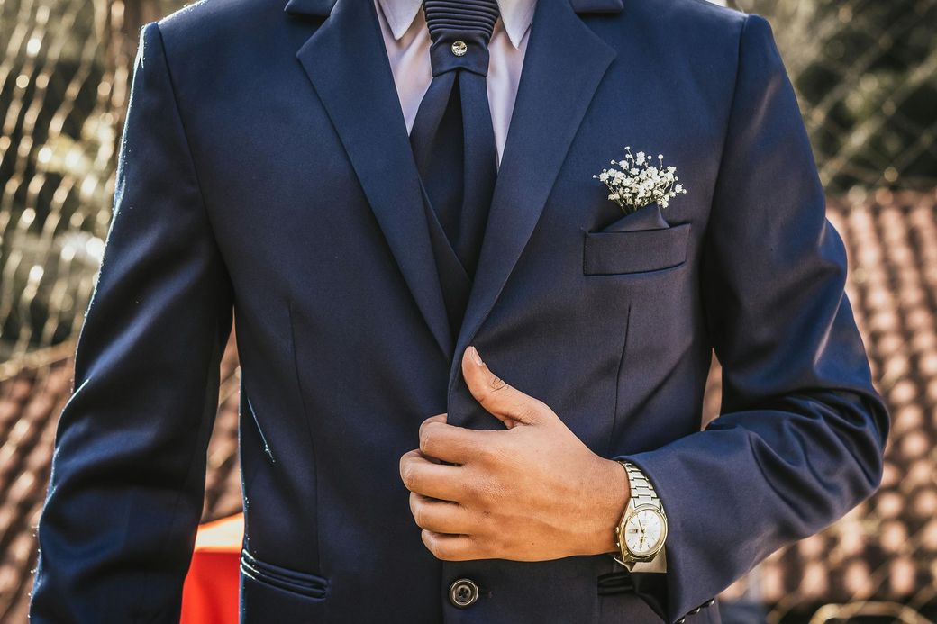 A person in a navy blue suit and vest with a floral boutonniere on the chest, wearing a silver watch on their wrist.