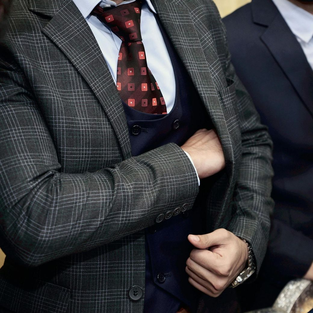 Person in a dark grey plaid blazer, blue vest, light blue shirt, and dark patterned tie with one hand tucked in a pocket.
