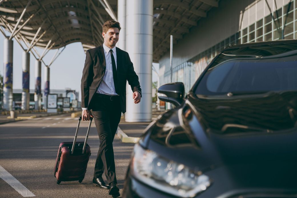 A person in a suit walks with a rolling suitcase toward a car parked at an airport terminal.