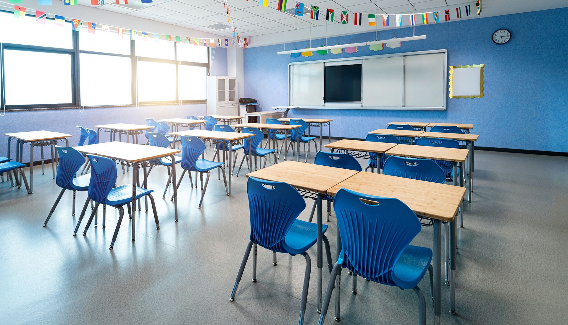 Empty classroom with blue walls, desks, chairs, and whiteboard. Sunlight streams in from the windows.
