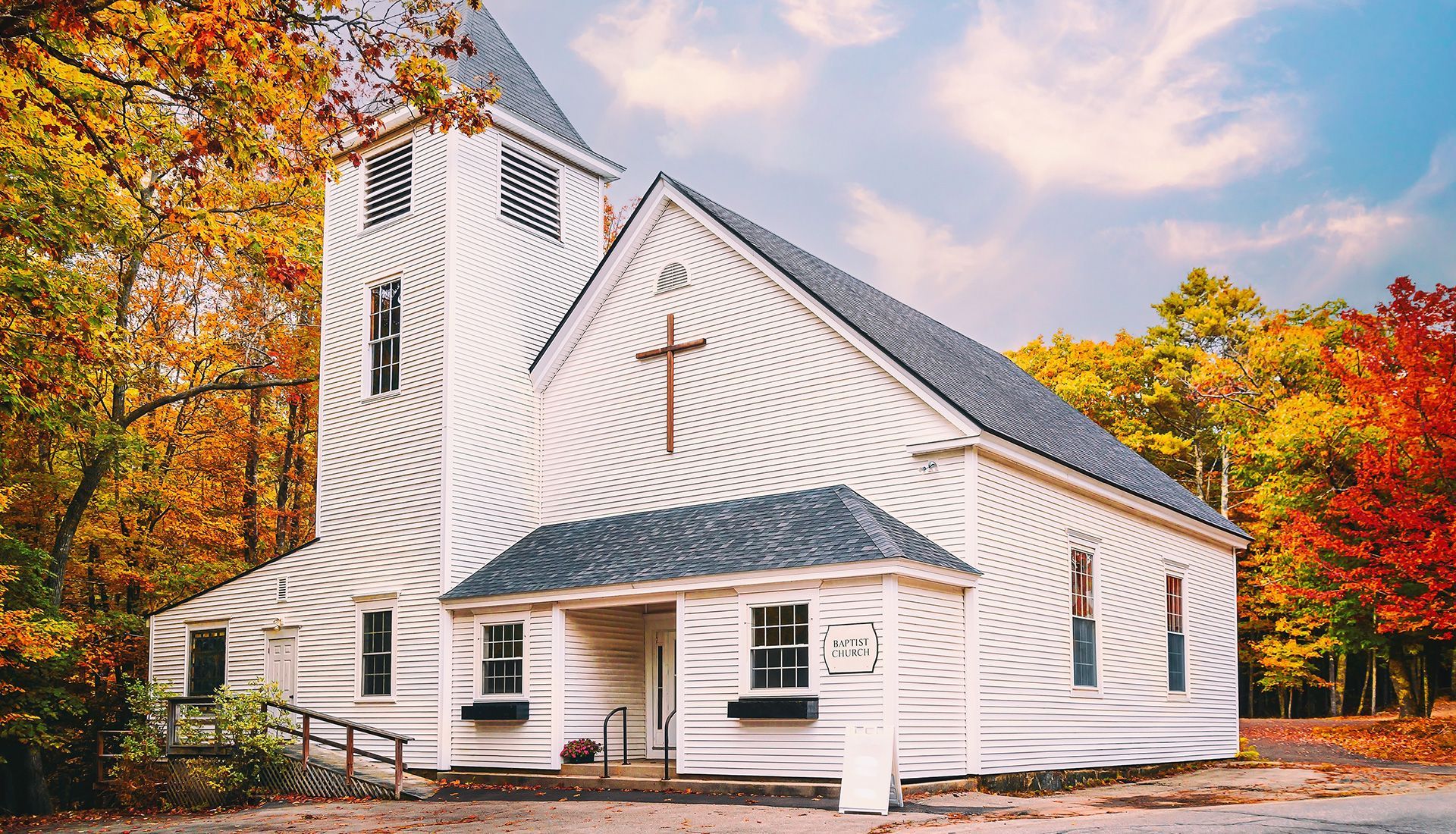 White church with cross, surrounded by fall foliage, under a cloudy sky.