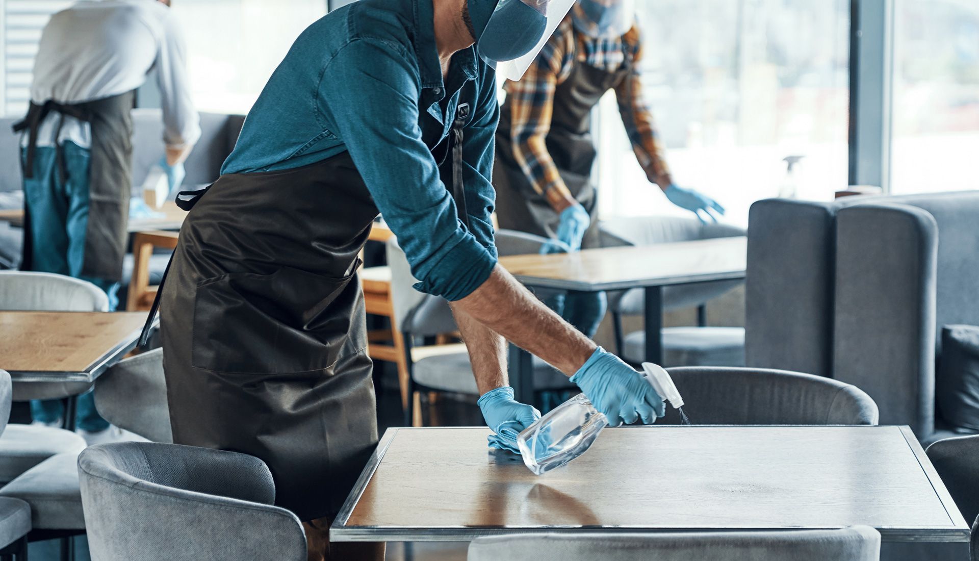 Staff sanitizing tables in a restaurant; wearing masks, gloves, and aprons.