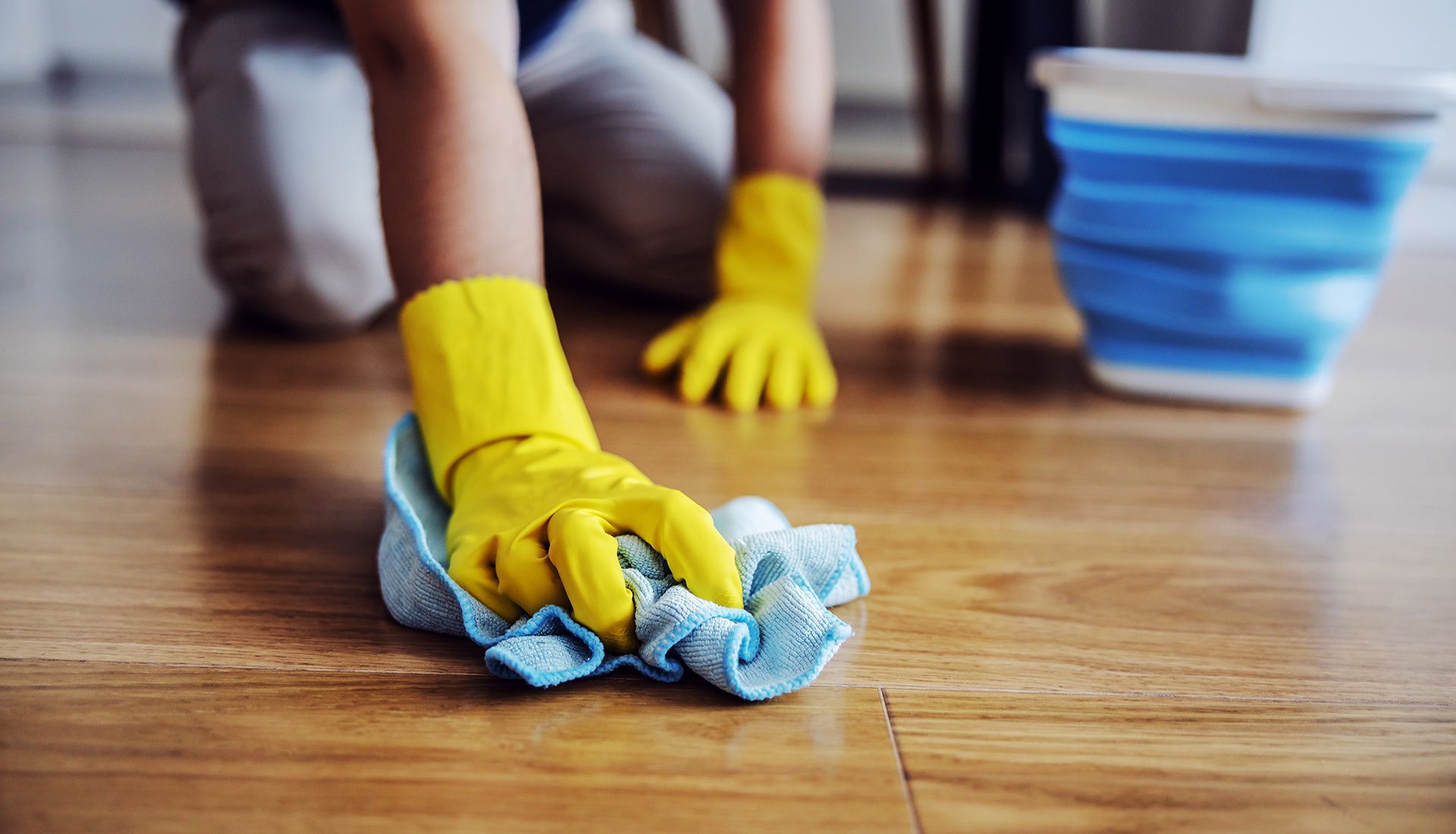 Person wearing yellow gloves, cleaning a wooden floor with a blue cloth and bucket.