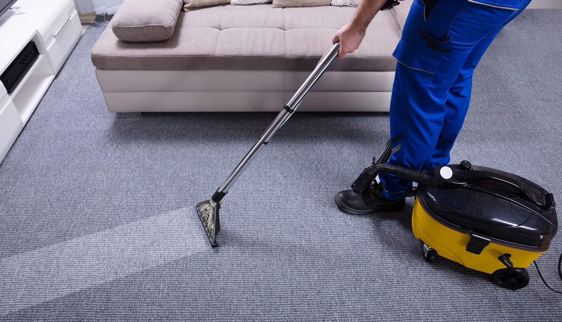 Person in blue uniform vacuuming a gray carpet near a beige couch. Yellow and black vacuum cleaner is on the floor.
