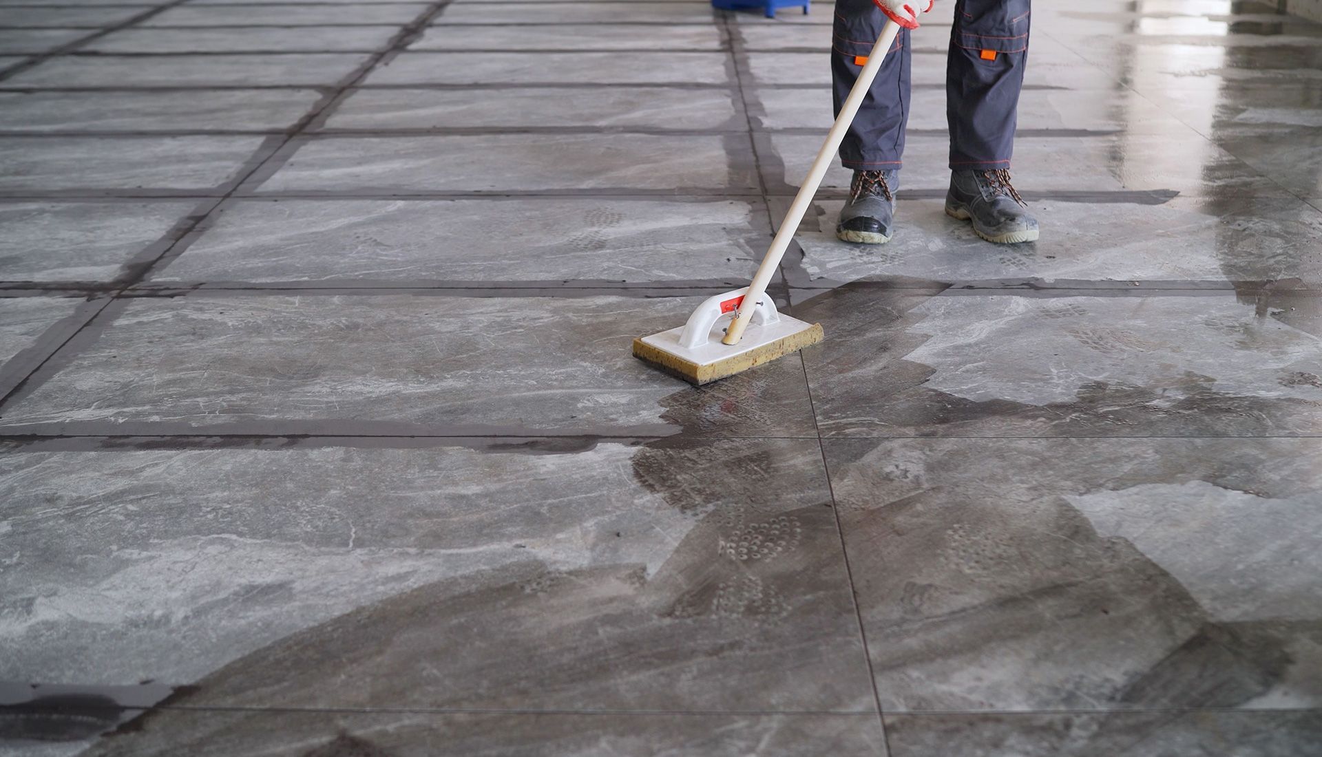 Person mopping a wet concrete floor in a building. Dark grout lines divide the floor.