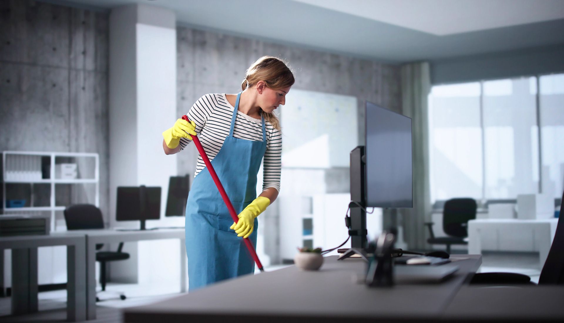 Woman mopping an office floor with a red handled mop. She wears an apron and yellow gloves.