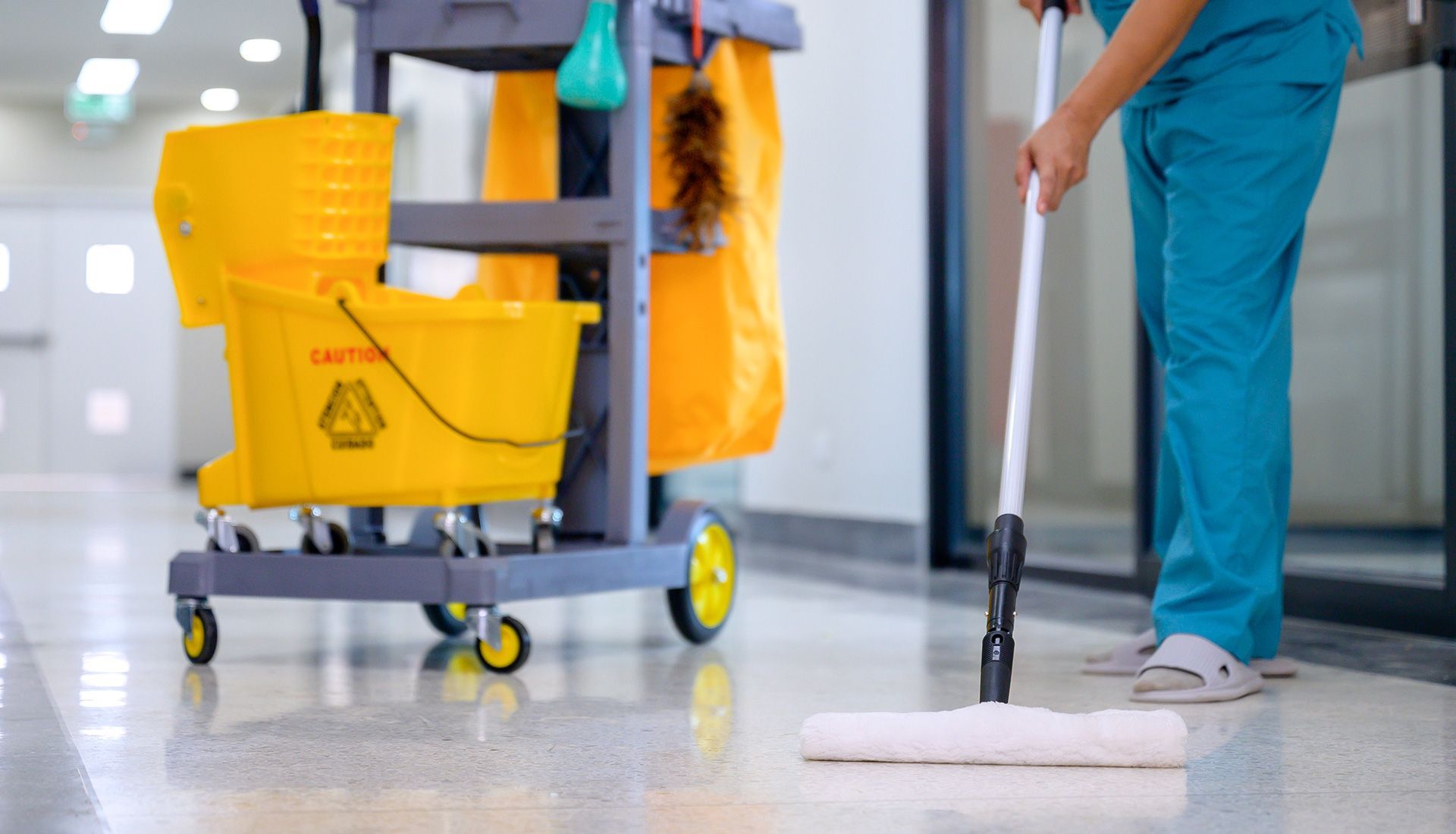 A person in teal scrubs mops a shiny floor next to a yellow cleaning cart in a hallway.