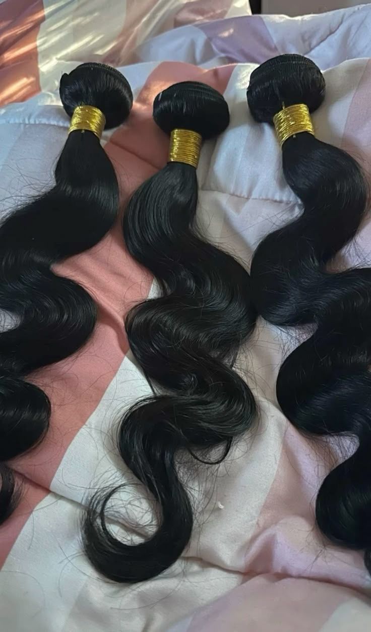Three black wavy hair bundles with gold bands on a pink satin surface