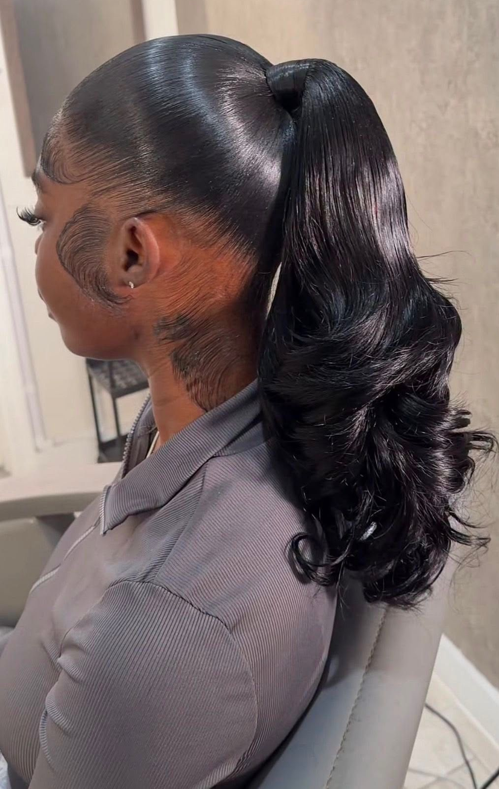Side-profile portrait of a woman with a sleek high ponytail, seated indoors against a neutral wall.