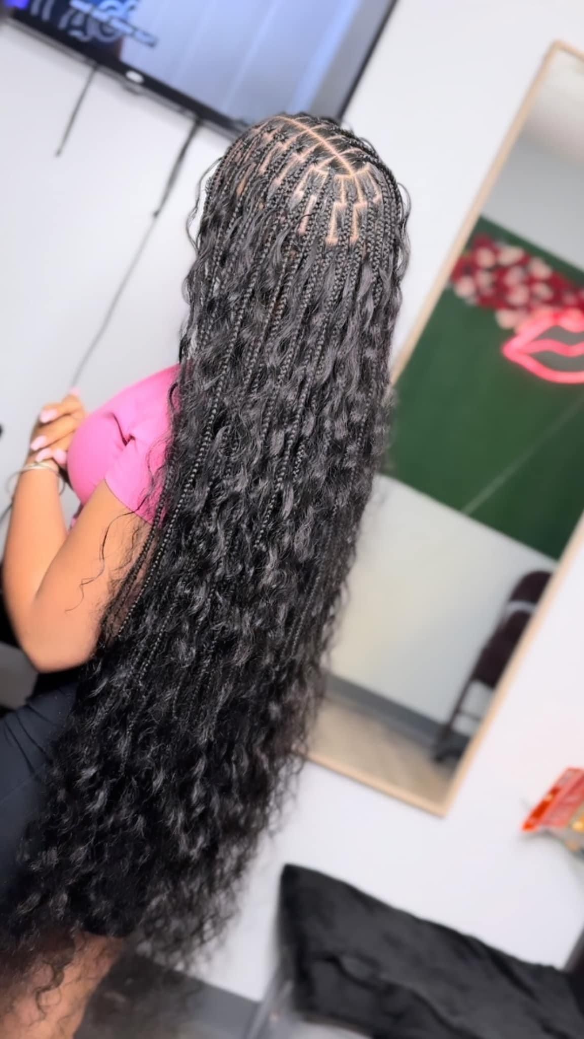 Woman with long black braids seated indoors, seen from behind in a pink top.