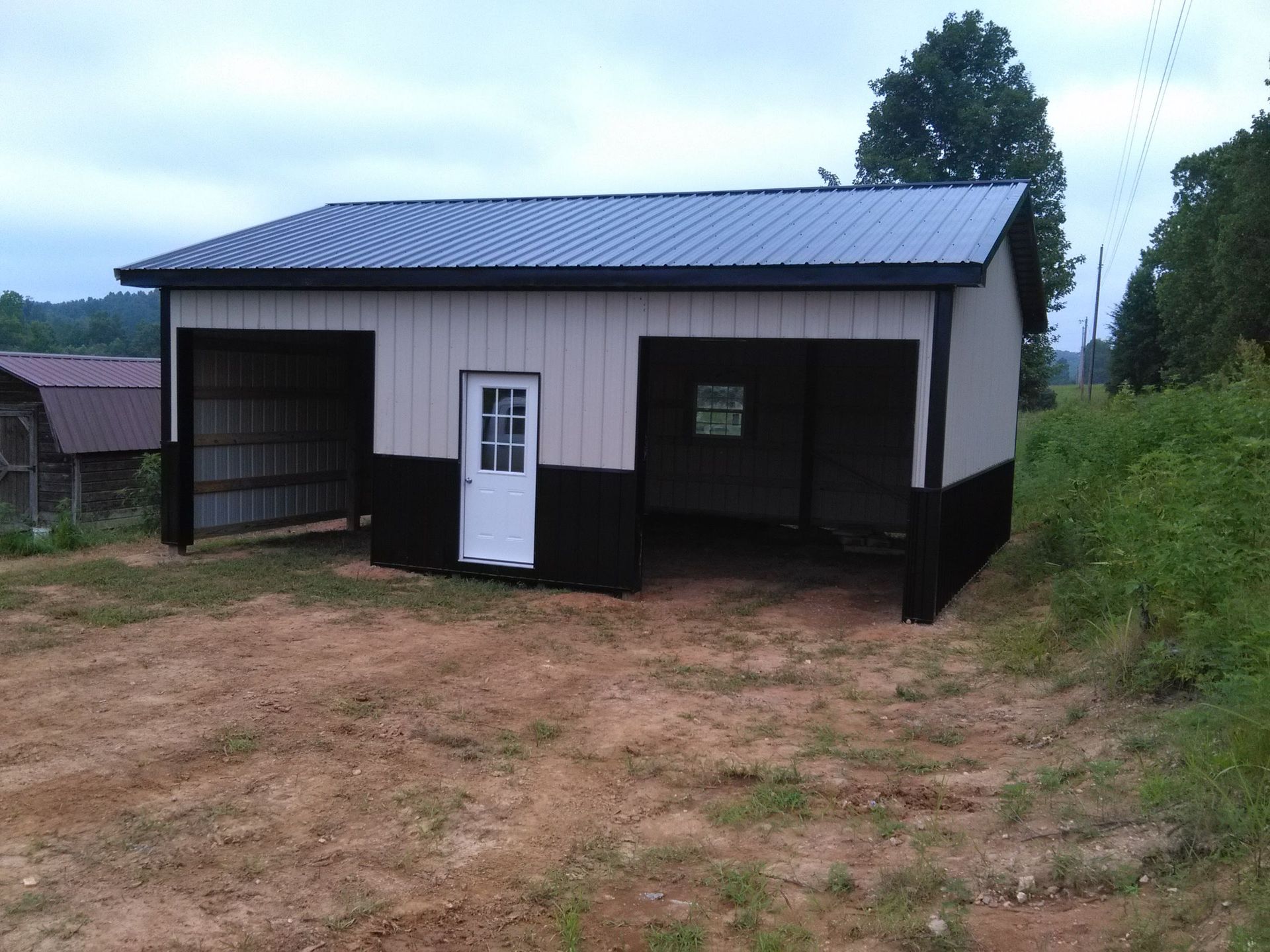 A garage with a black roof and a white door is sitting in the middle of a dirt field.