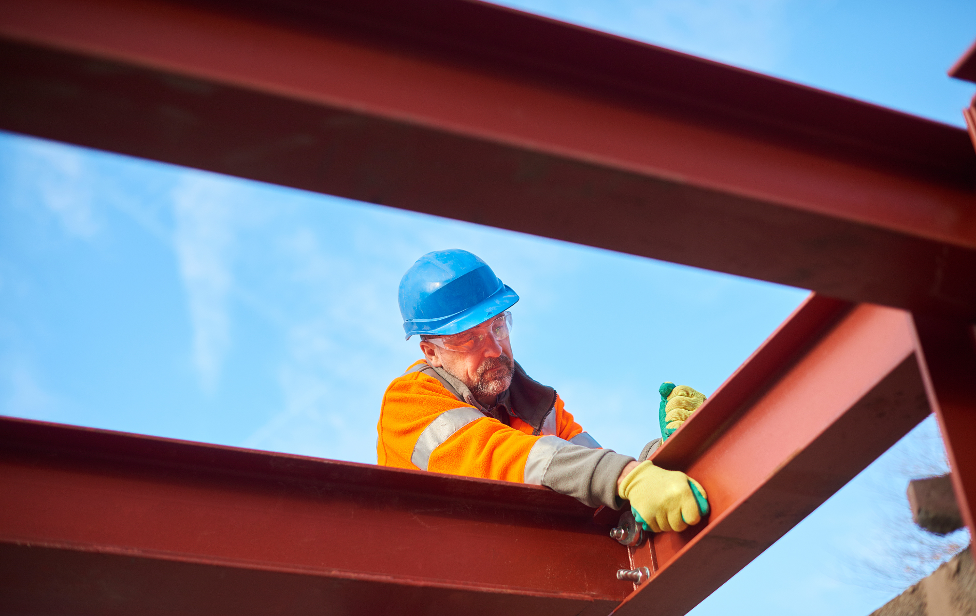 Construction worker in blue hard hat and orange vest working on red metal beams under blue sky.