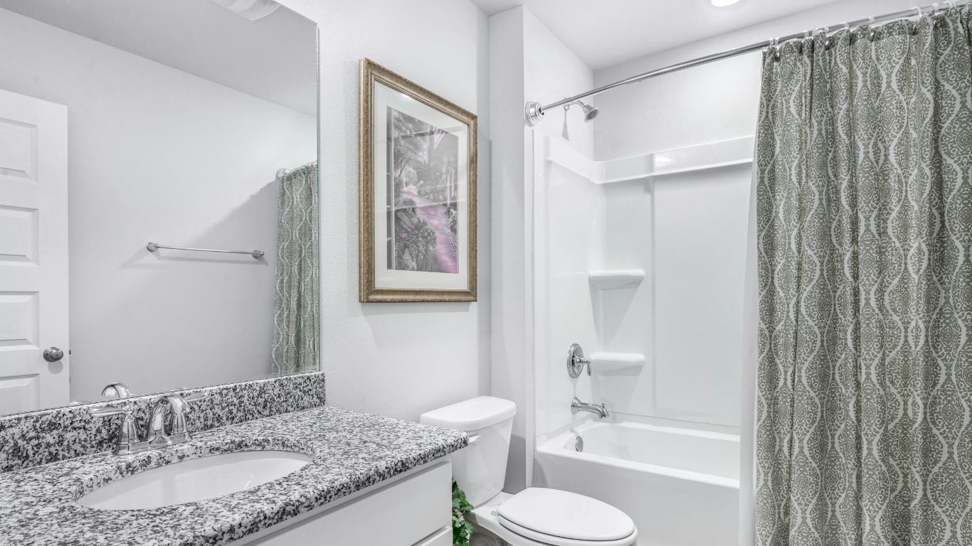 A modern bathroom with a granite vanity, white toilet, and a white bathtub enclosed by a patterned grey shower curtain.