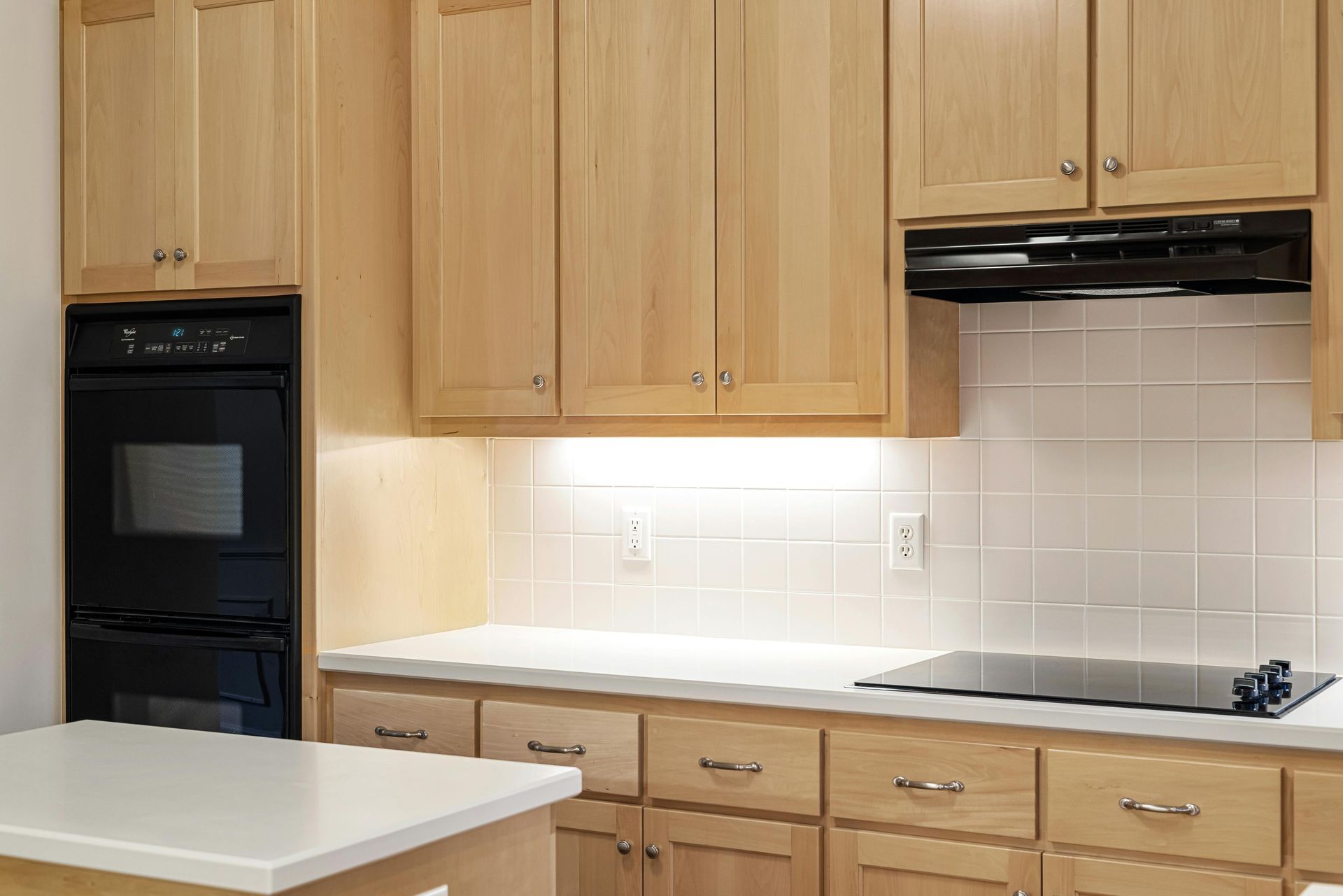A modern kitchen with light wood cabinets, white countertops, a black double oven, and a glass stovetop with a vent.
