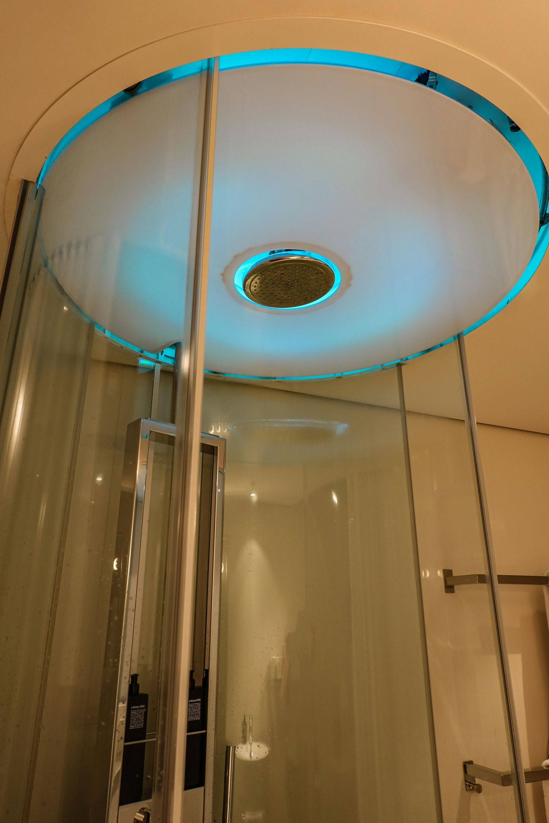 Modern shower with a circular ceiling light emitting a soft blue glow above a glass door and chrome fixtures.