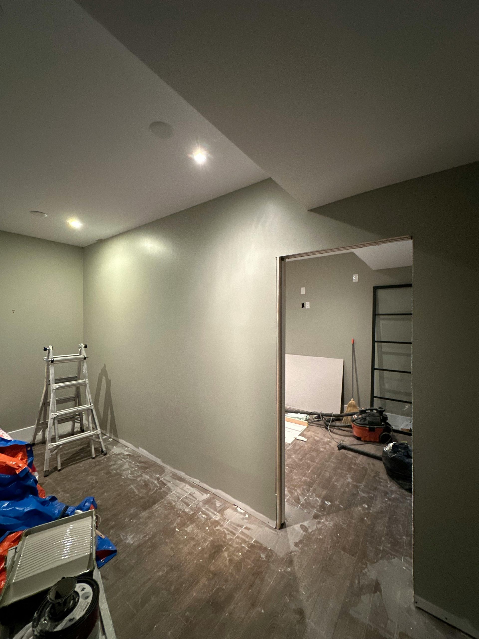 A room with freshly painted sage-green walls, a metal ladder, and an open doorway leading to an unfinished room.