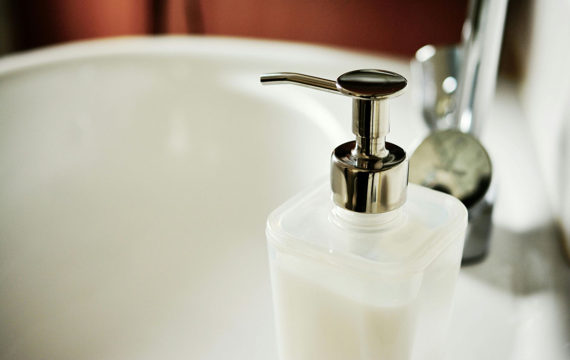 A white, rectangular soap dispenser with a metallic pump sitting next to a chrome faucet in a bathroom sink.