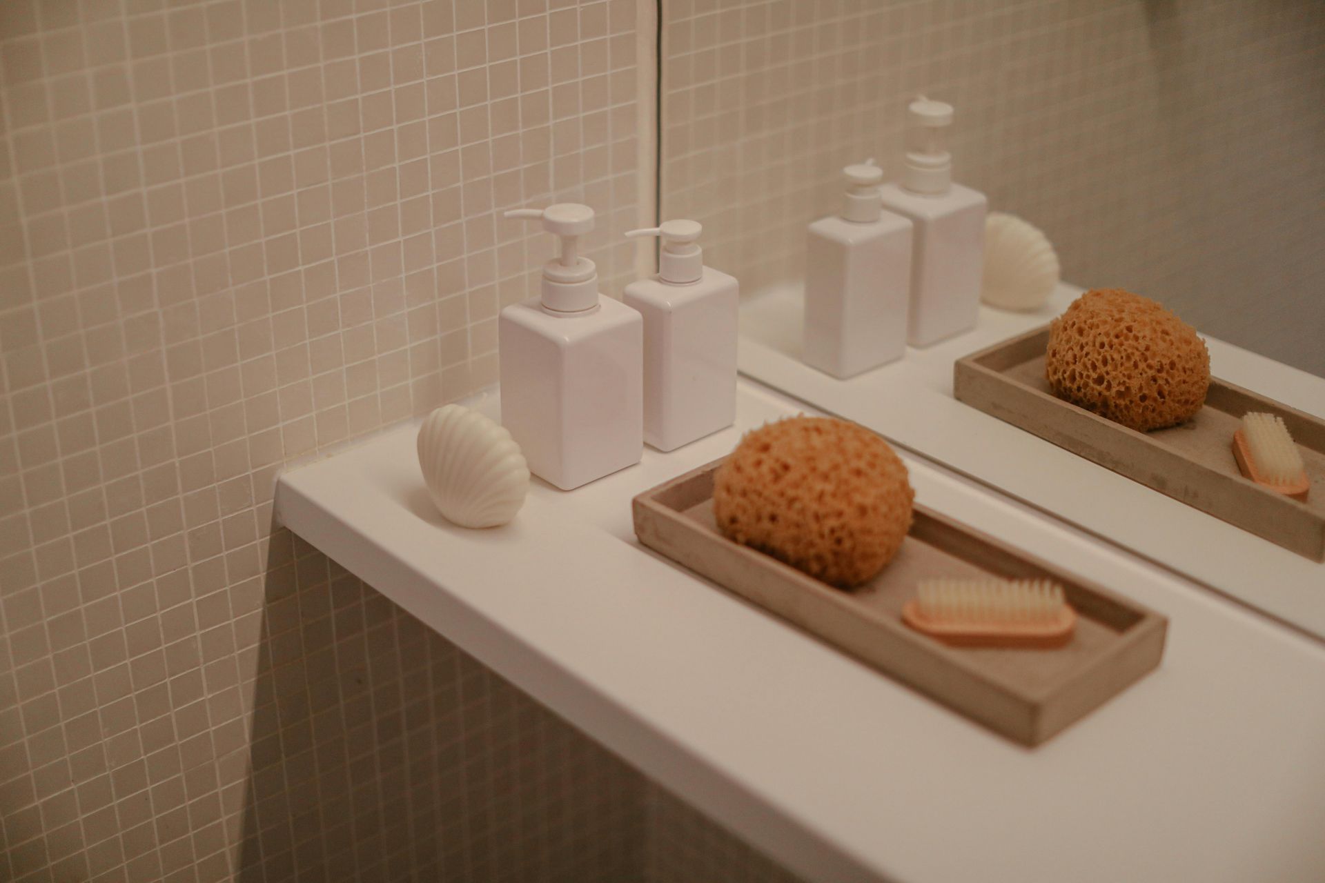 Minimalist bathroom counter with two white soap dispensers, a natural sponge, and a small brush on a tray by a mirror.