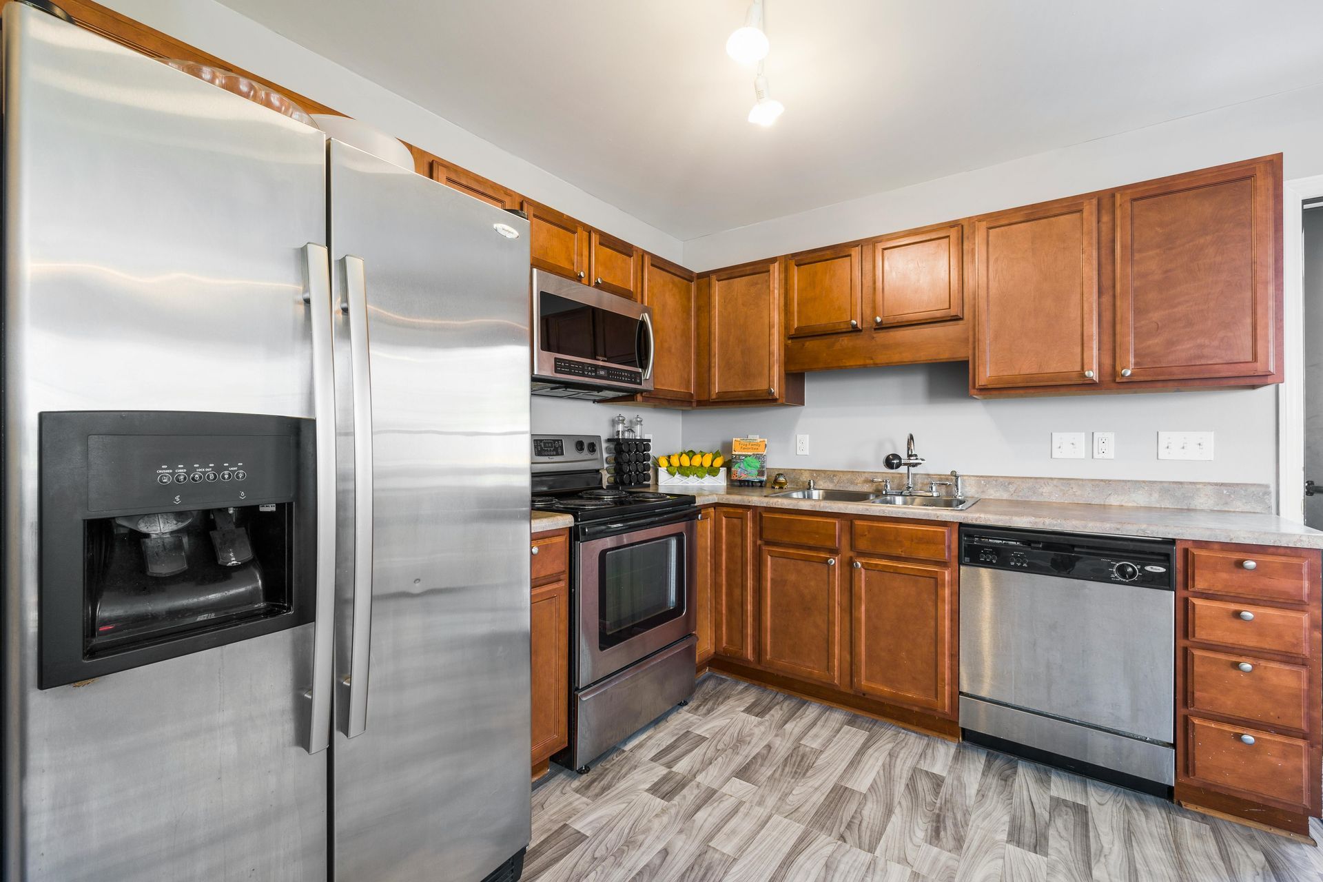 A kitchen with stainless steel appliances, brown wooden cabinets, gray countertops, and light-toned wood-grain flooring.
