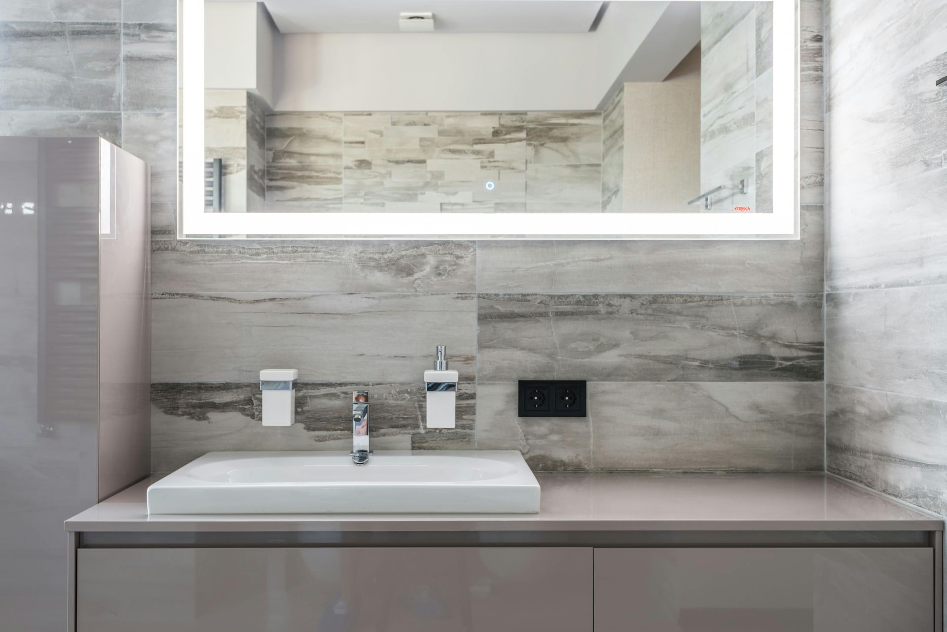 A modern bathroom vanity with a white rectangular sink, light grey stone-textured walls, and an illuminated mirror.