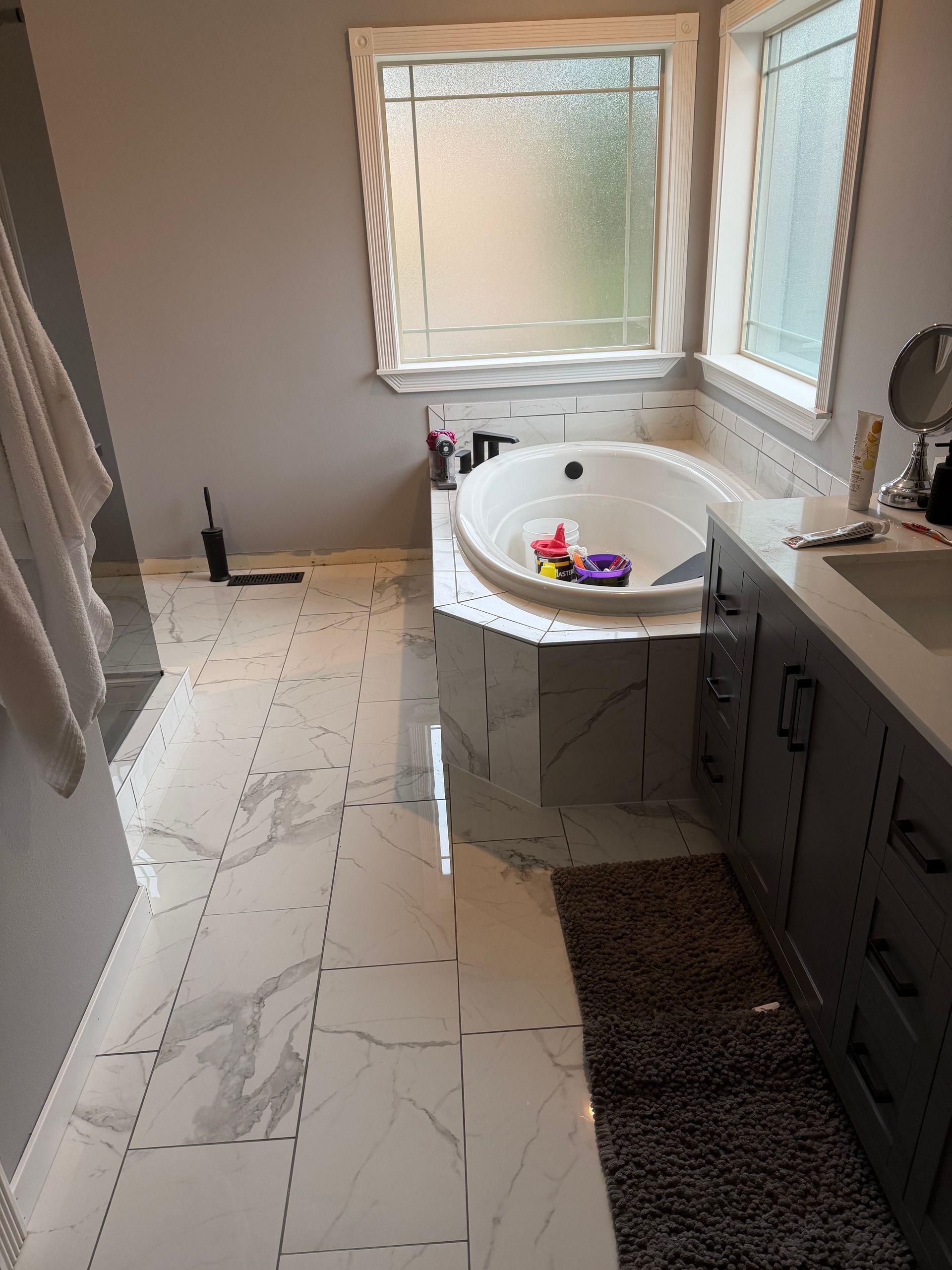 Bathroom with white marble floor and tub, gray cabinets, and frosted window.