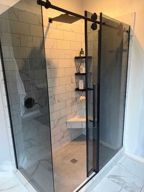 A modern shower with sliding glass door, black hardware, and gray tile. Includes a built-in seat and shelves.