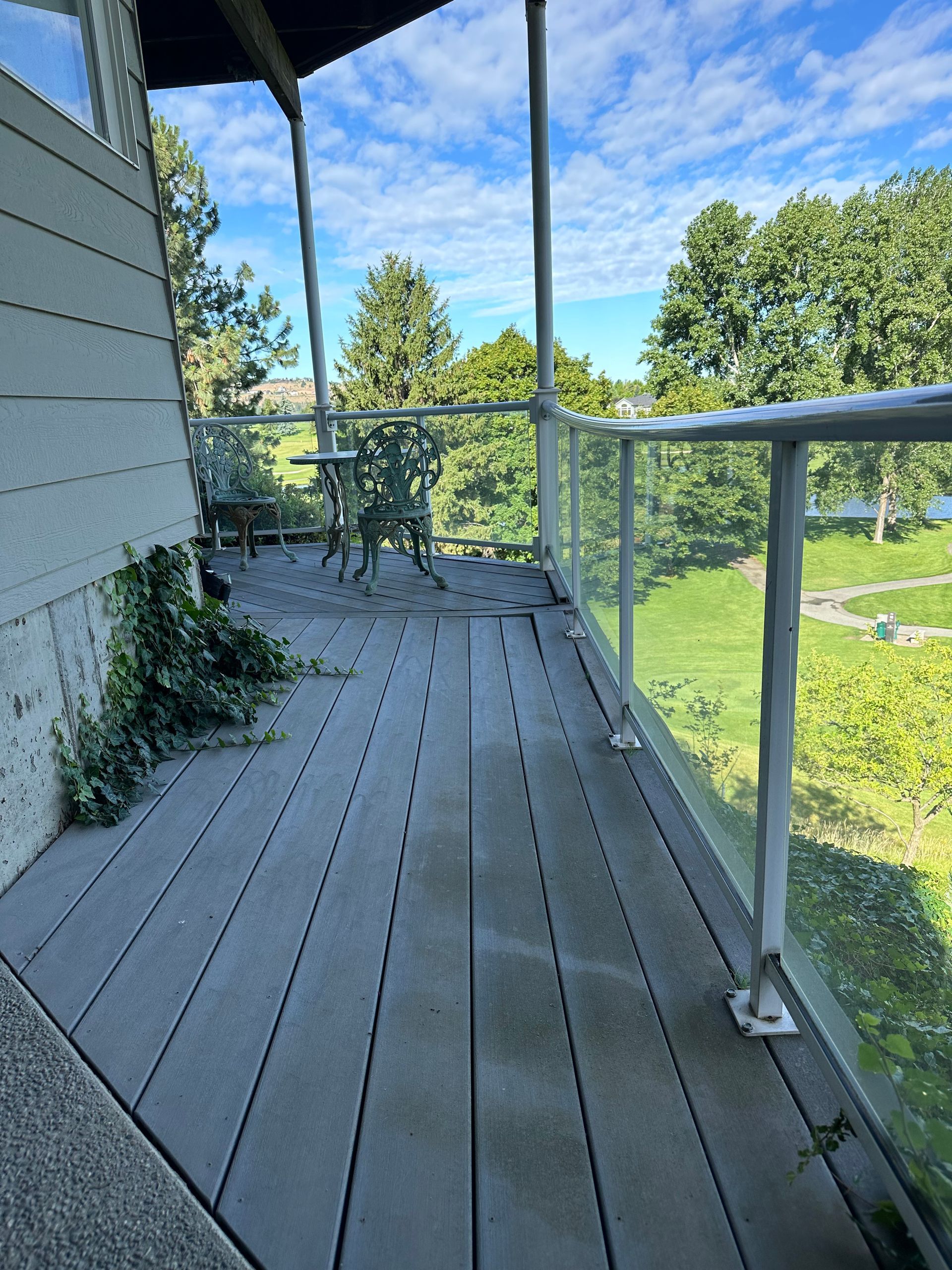 Outdoor deck with glass railing and gray composite decking; trees and a blue sky are in the background.