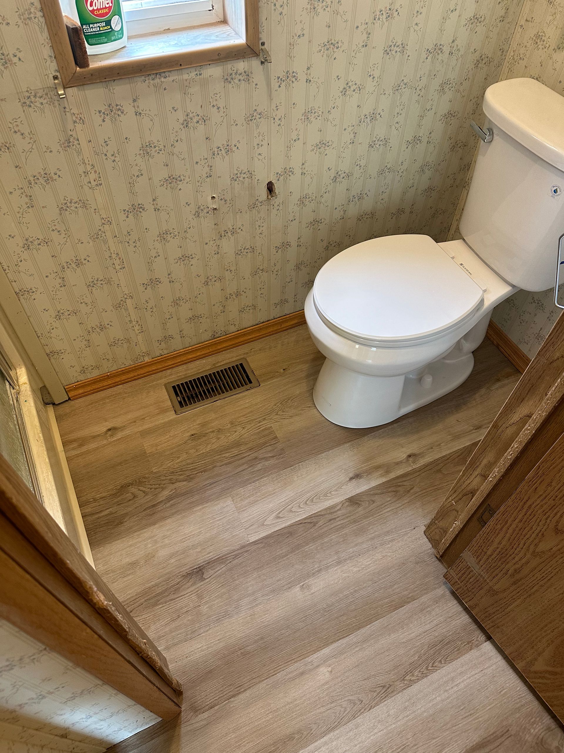 Bathroom with a toilet, tan wood-look floor, and patterned wallpaper.