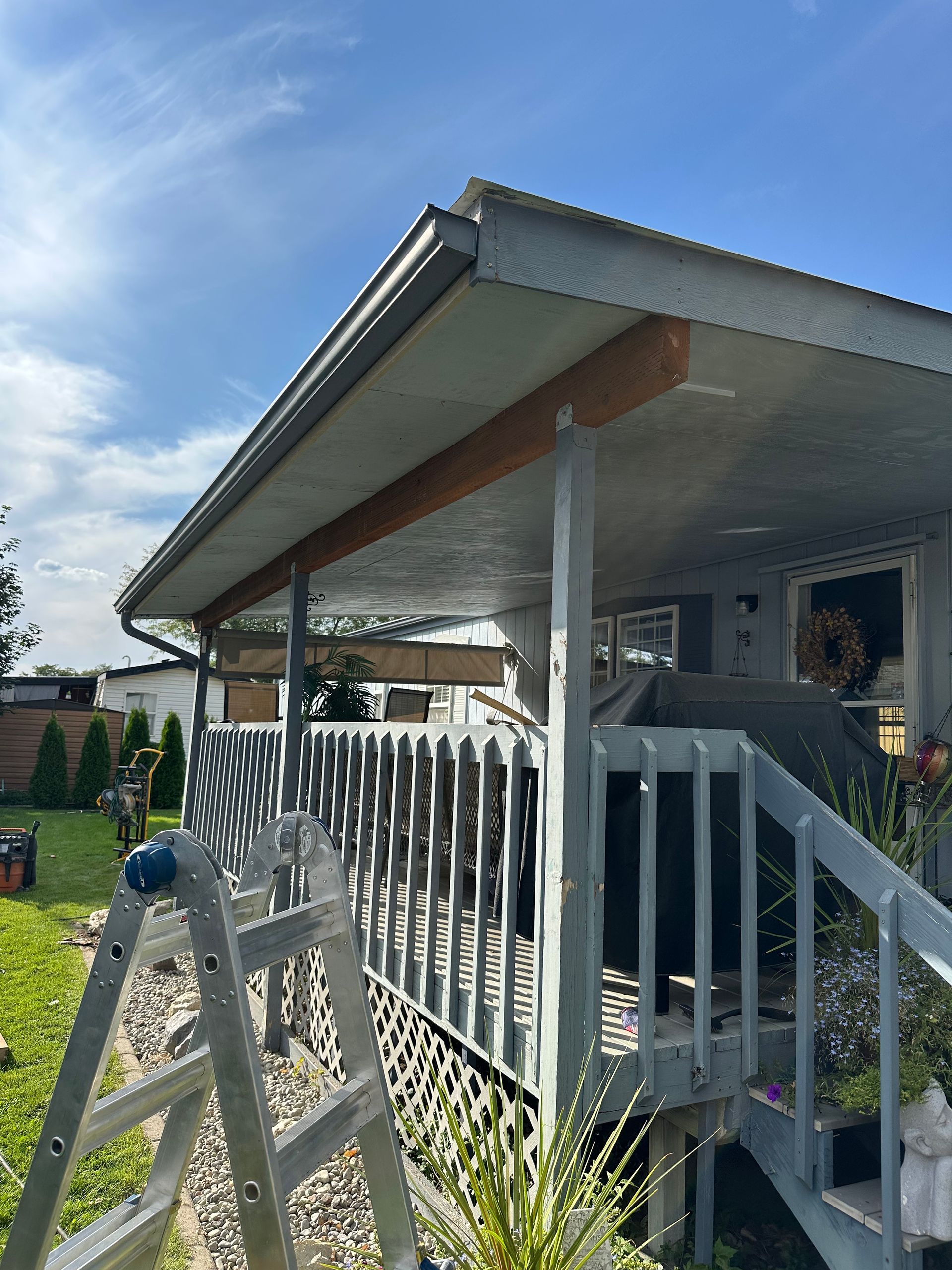 A porch with a gray railing, wooden supports, and a ladder on the grass under a blue sky.