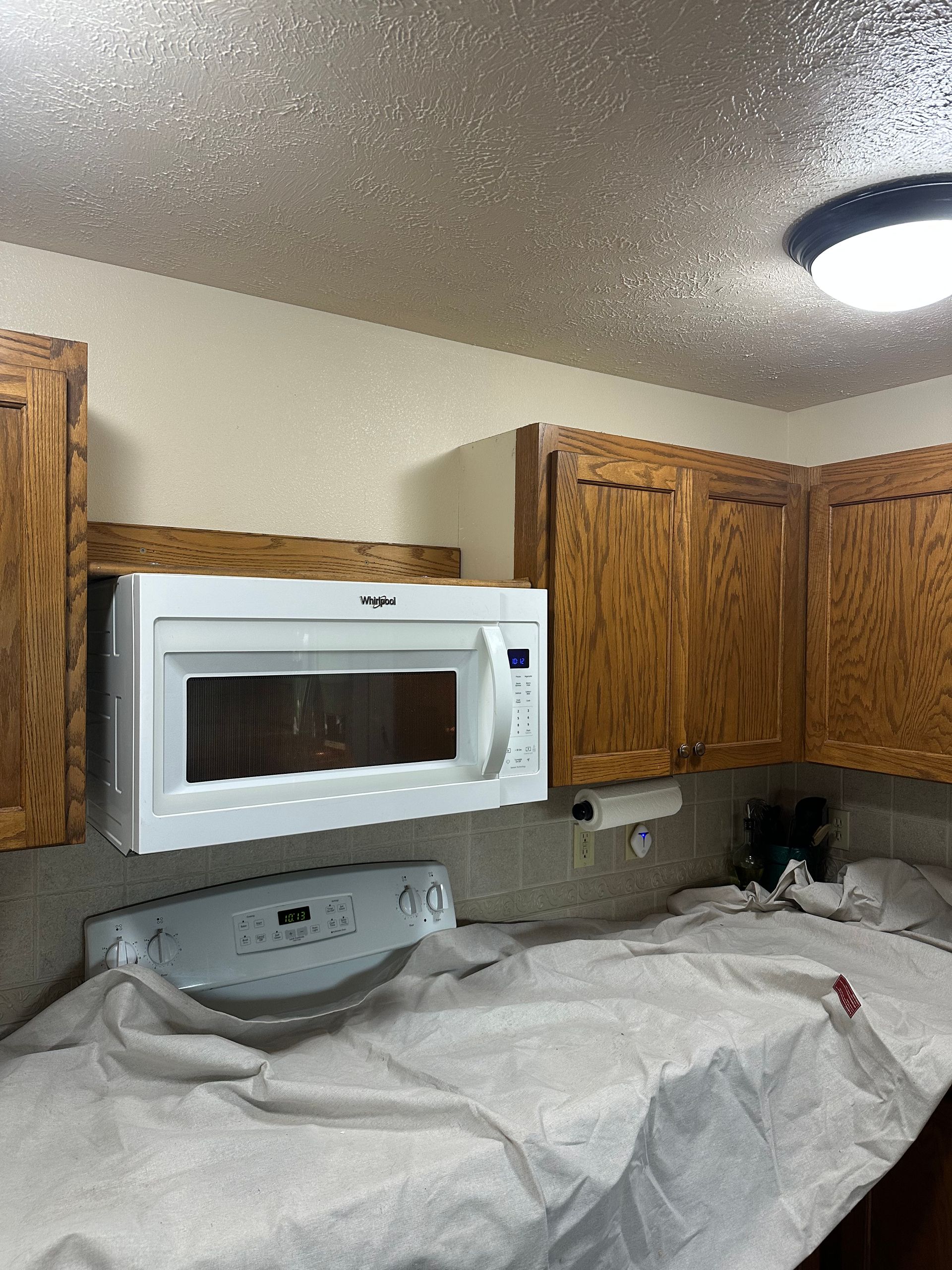 Kitchen with wooden cabinets, a white microwave, stove, and covered counters; painting in progress.