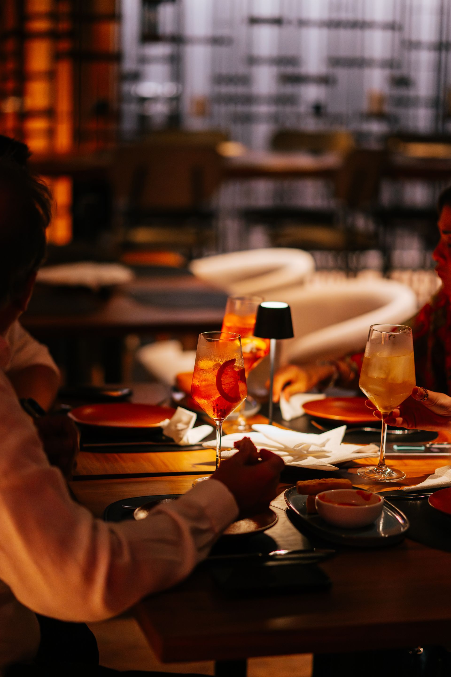 Personas cenando en un restaurante, bajo una luz tenue. Cócteles y platos en la mesa.