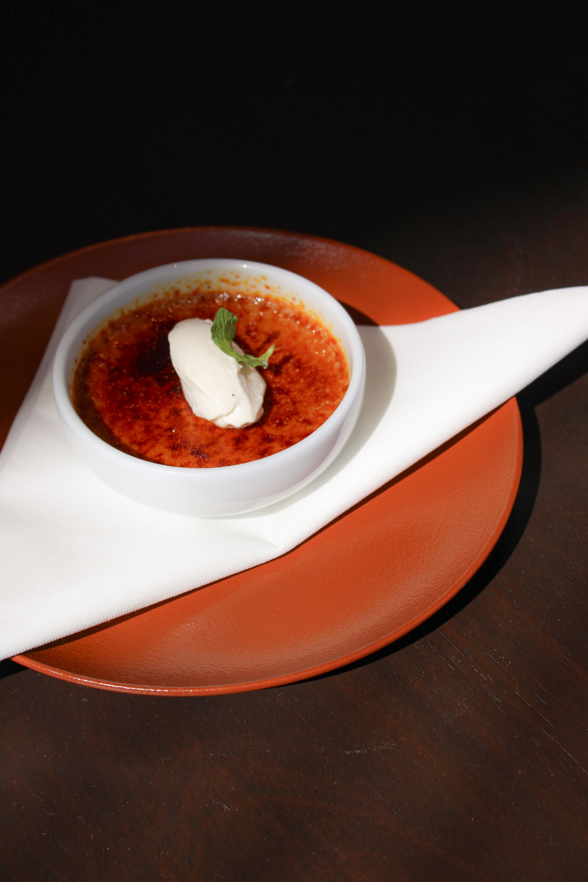 Crème brûlée en tazón blanco sobre un plato rojo con una servilleta blanca; cubierta con crema batida y una ramita de menta.