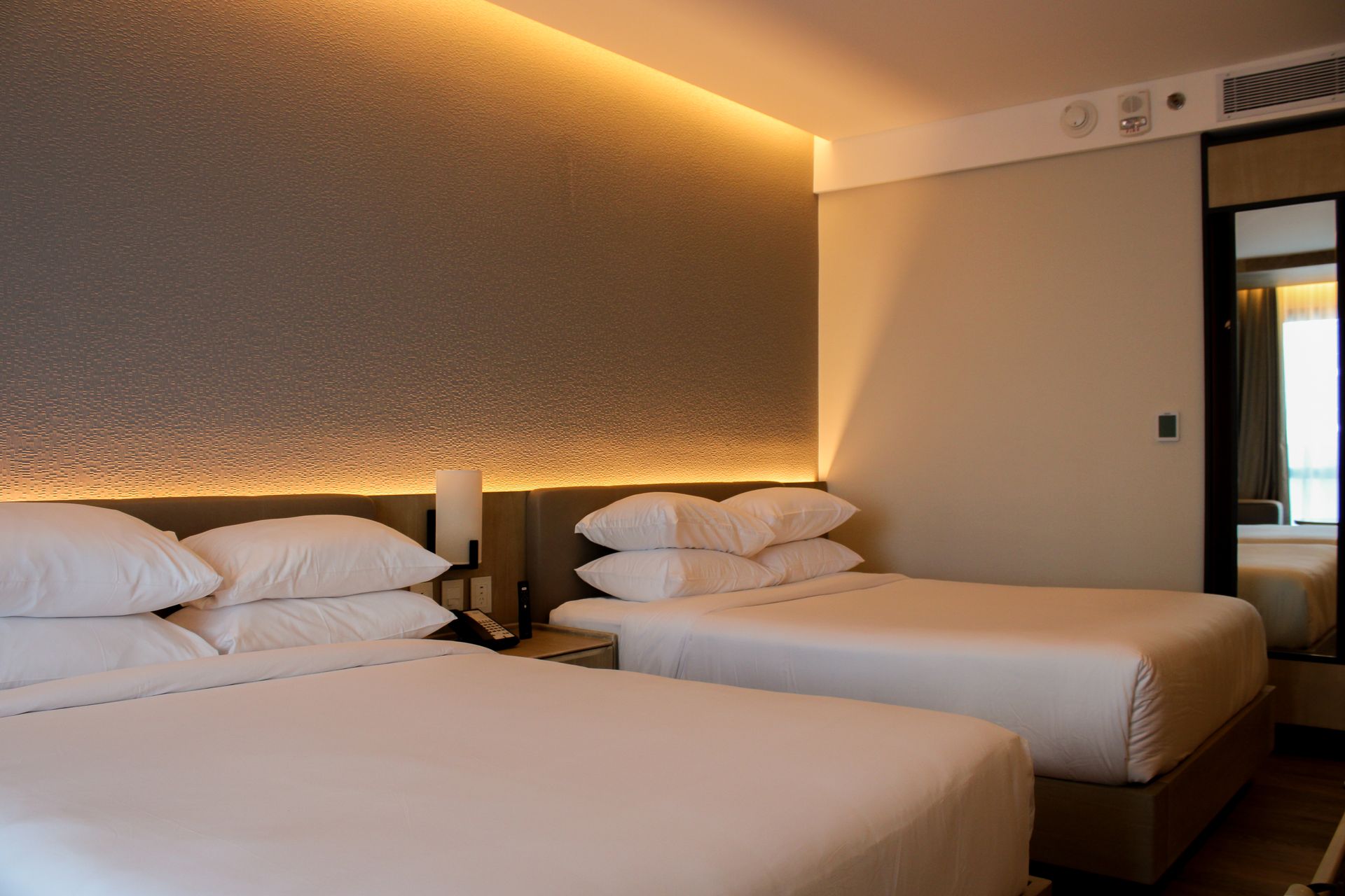 Habitación de hotel con dos camas, pared con acento gris texturizado con iluminación cálida sobre las camas, espejo a la derecha.