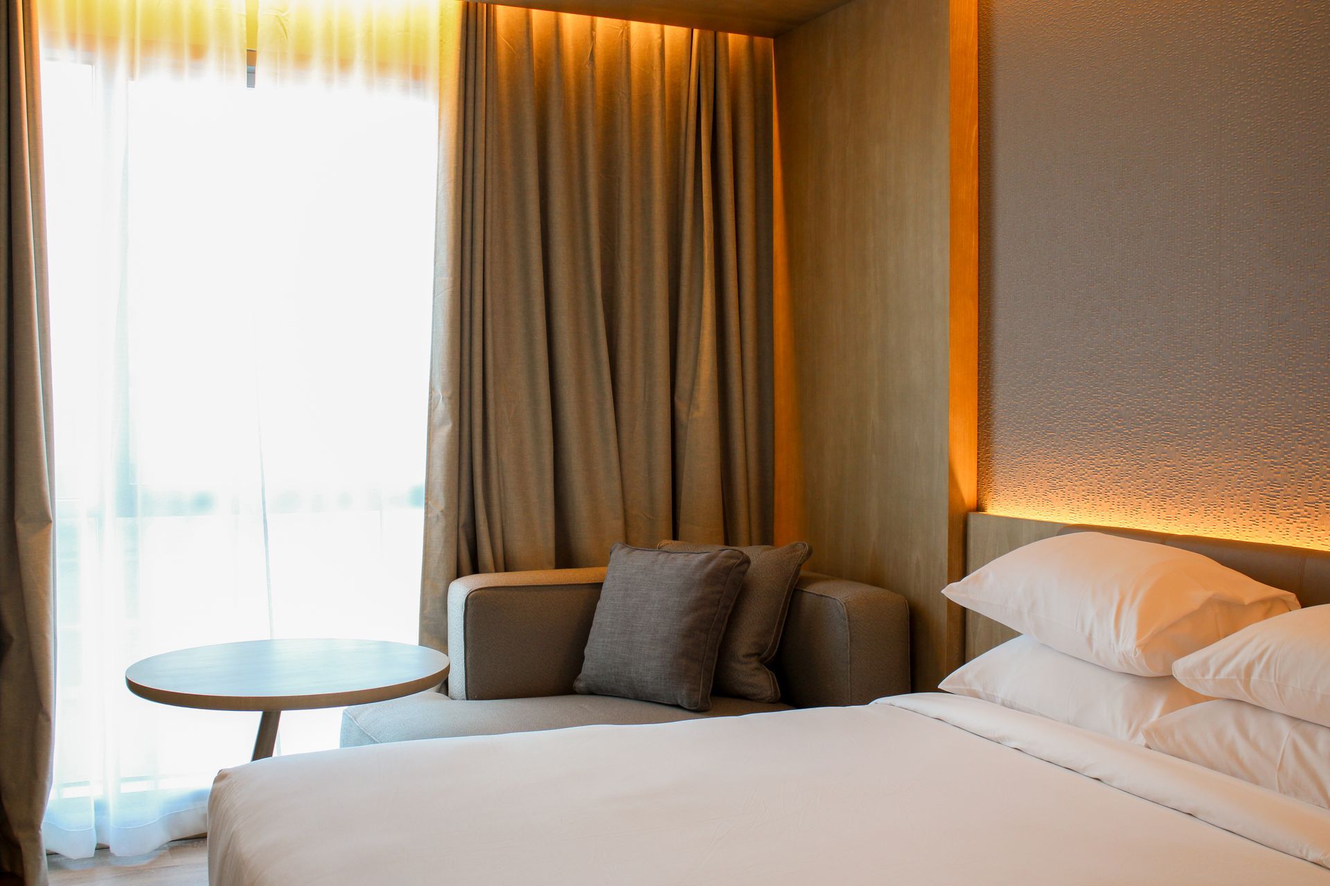 Habitación de hotel con cama, pequeño sofá, mesa redonda, ventana y paredes y cortinas en tonos neutros.