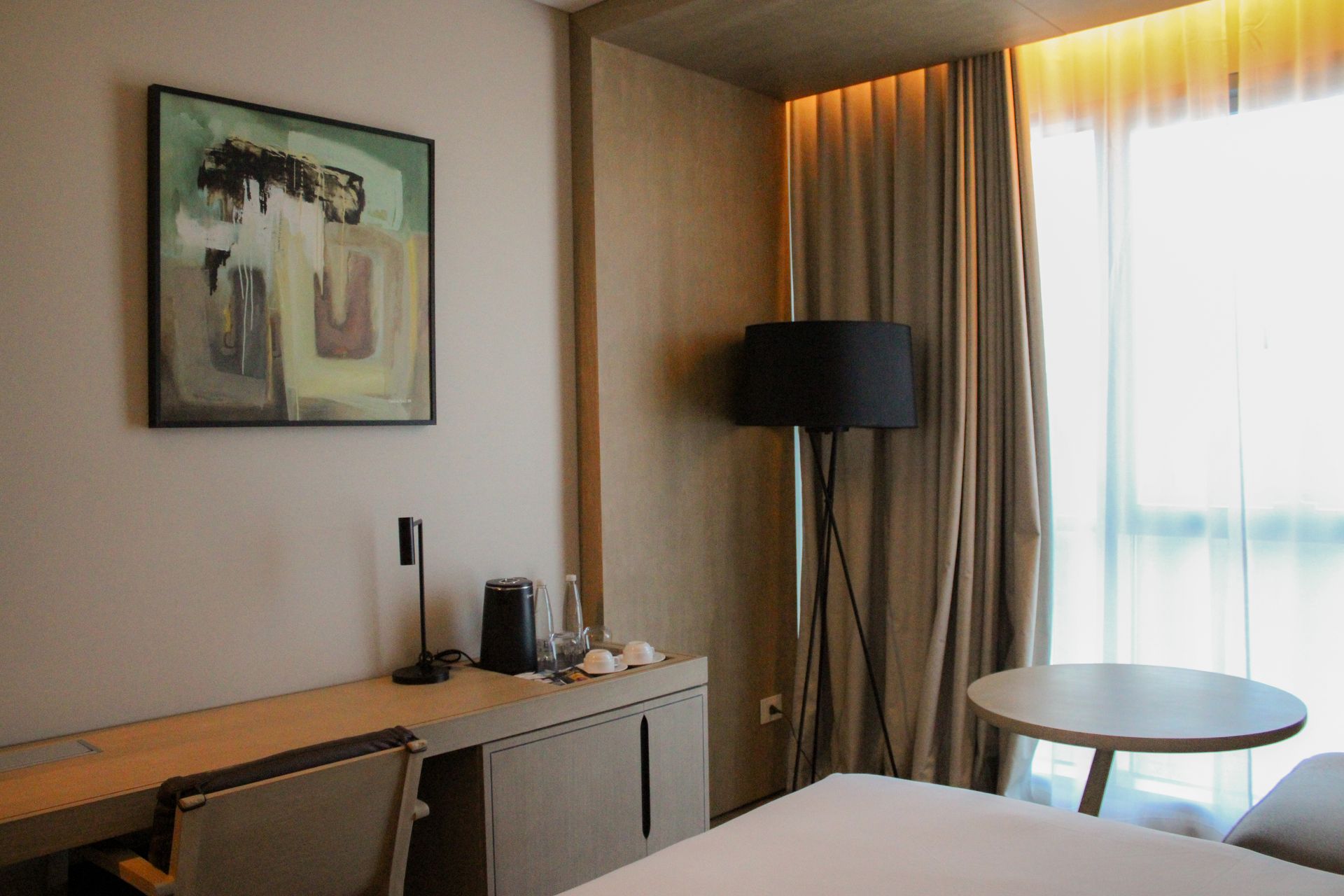 Interior de habitación de hotel: escritorio con silla, arte, lámpara de pie, cama, mesa redonda, cortinas y una ventana.