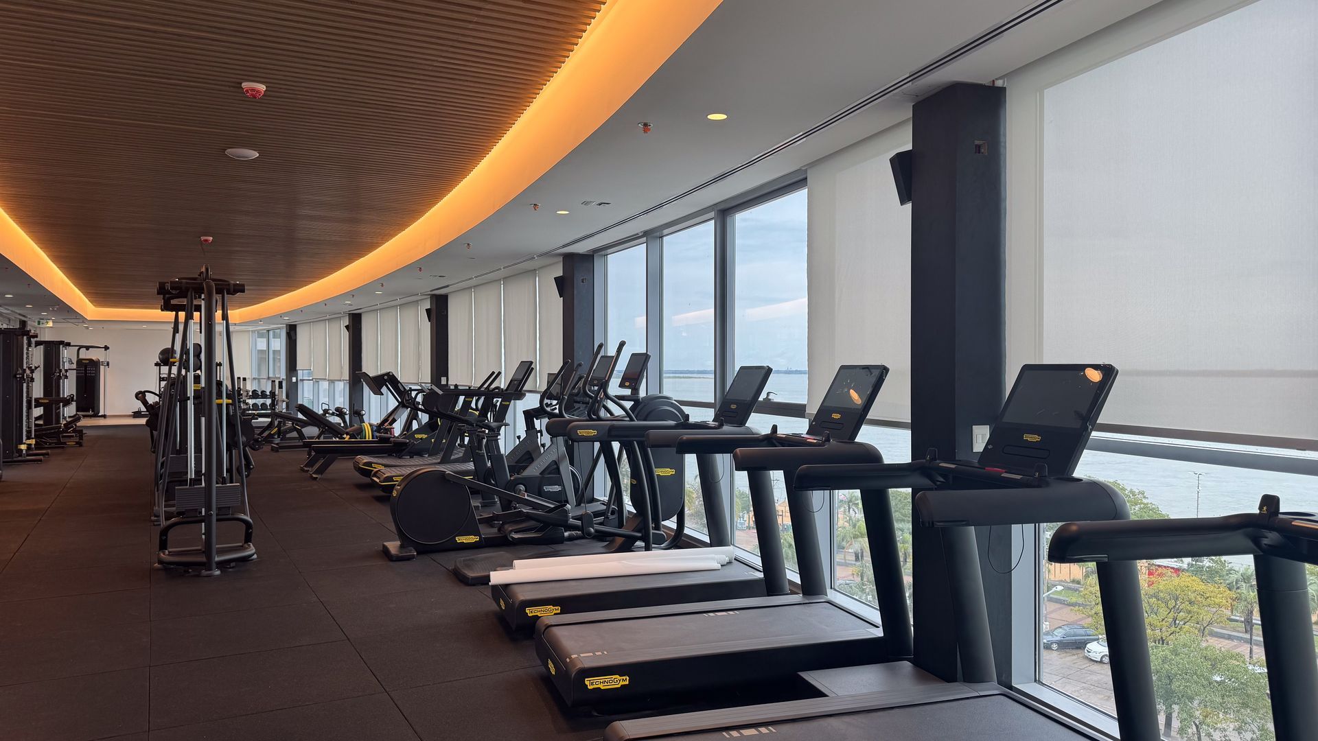 Gimnasio con cintas de correr, máquinas elípticas y equipos de pesas, junto a grandes ventanales con vista a un cuerpo de agua.