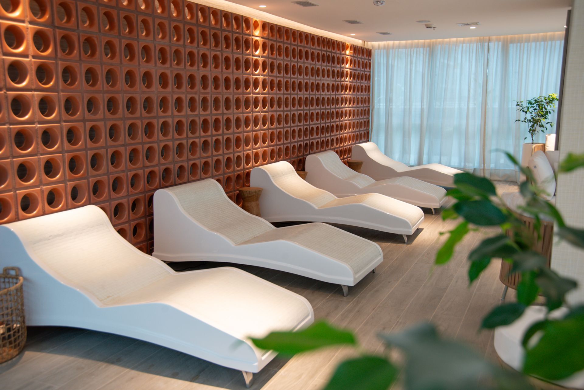 Sala de relajación con tumbonas blancas, pared de terracota texturizada y cortinas transparentes.
