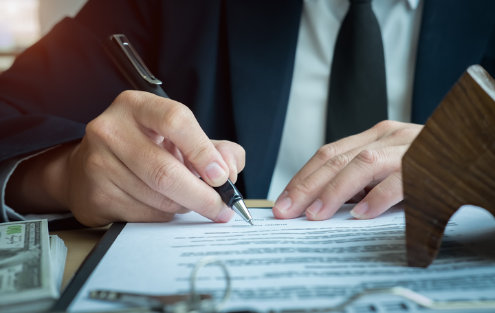 Hands signing a document on a desk with a pen, paperwork and a cash stack nearby