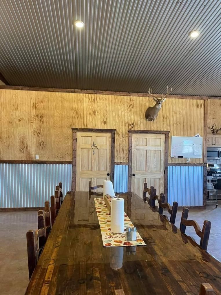 A long, polished wooden table in a rustic room with corrugated metal ceilings, wood-paneled walls, and two wooden doors.