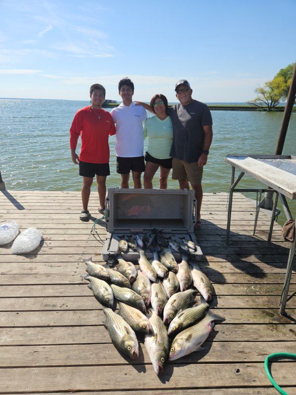 Four people stand on a wooden dock by the water, posing behind a large cooler filled with their catch of many fish.