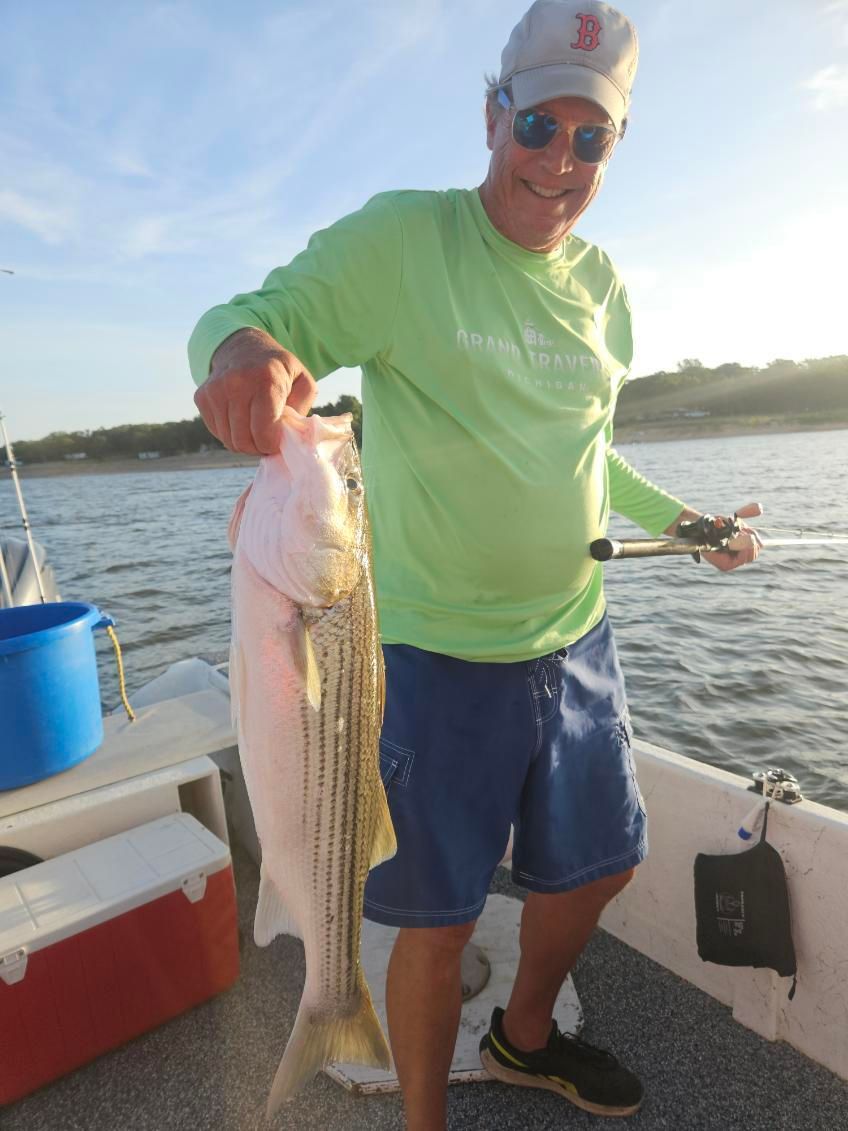 A person in a neon green shirt and sunglasses holding a striped bass while standing on a boat on the water.