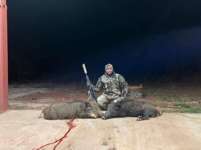 A person in camouflage gear holds a rifle while kneeling behind two harvested wild boars on a concrete surface at night.