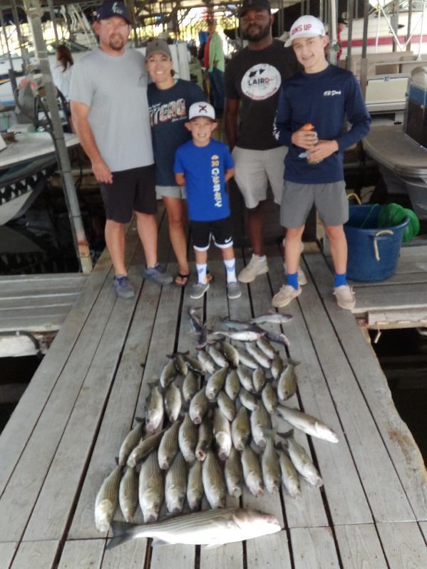 A group of people pose on a wooden dock with a large catch of fish laid out in front of them at a boat marina.