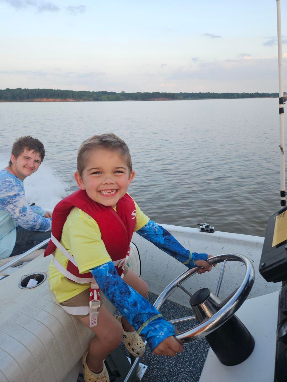 A child wearing a yellow shirt and red life jacket smiles while steering a boat on a calm body of water near a person.