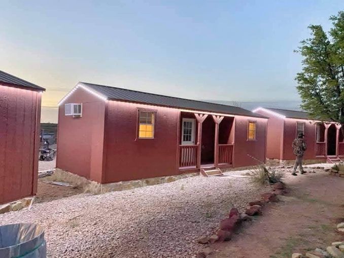 Rows of small red cabins with covered porches and illuminated rooflines stand on a gravel path at dusk.