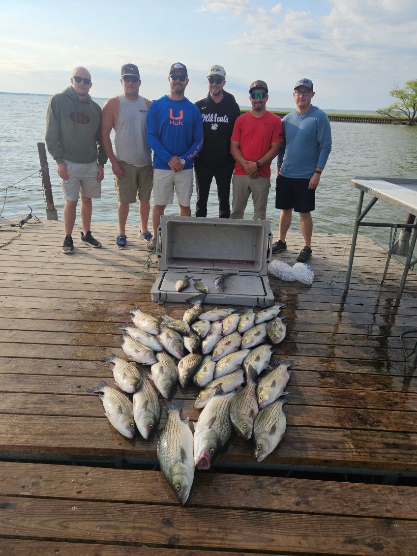 Six people stand on a wooden dock by a lake behind a large catch of fish spread out for display.