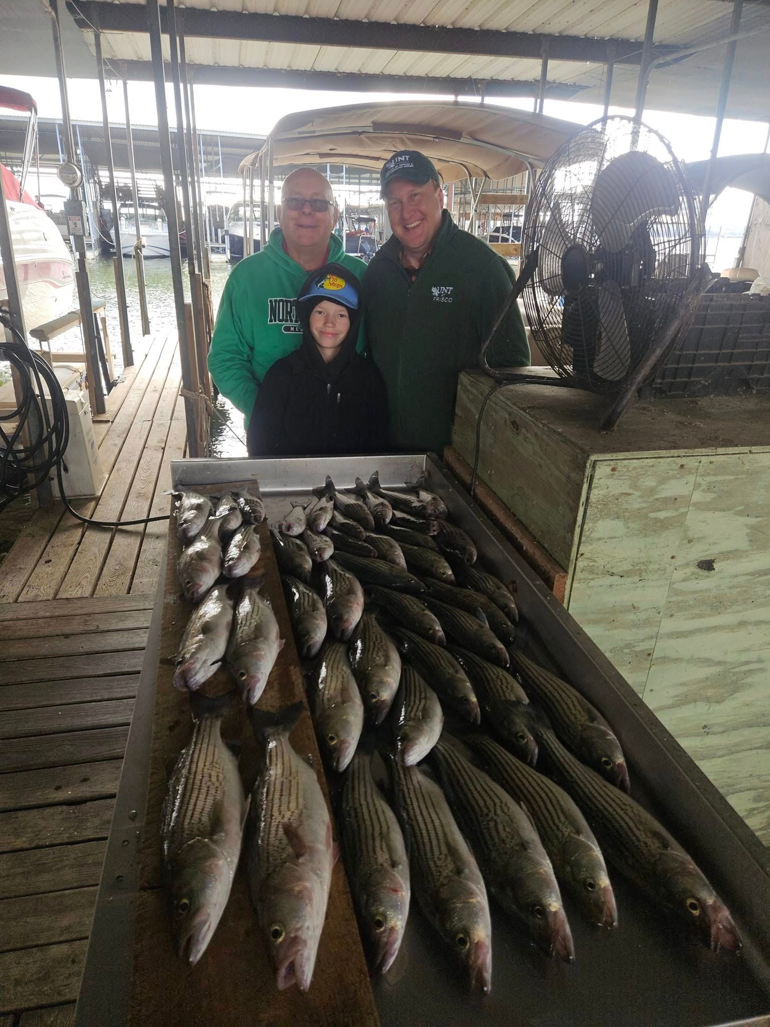 Three people stand behind a long wooden table filled with many freshly caught striped bass at a covered boat dock.