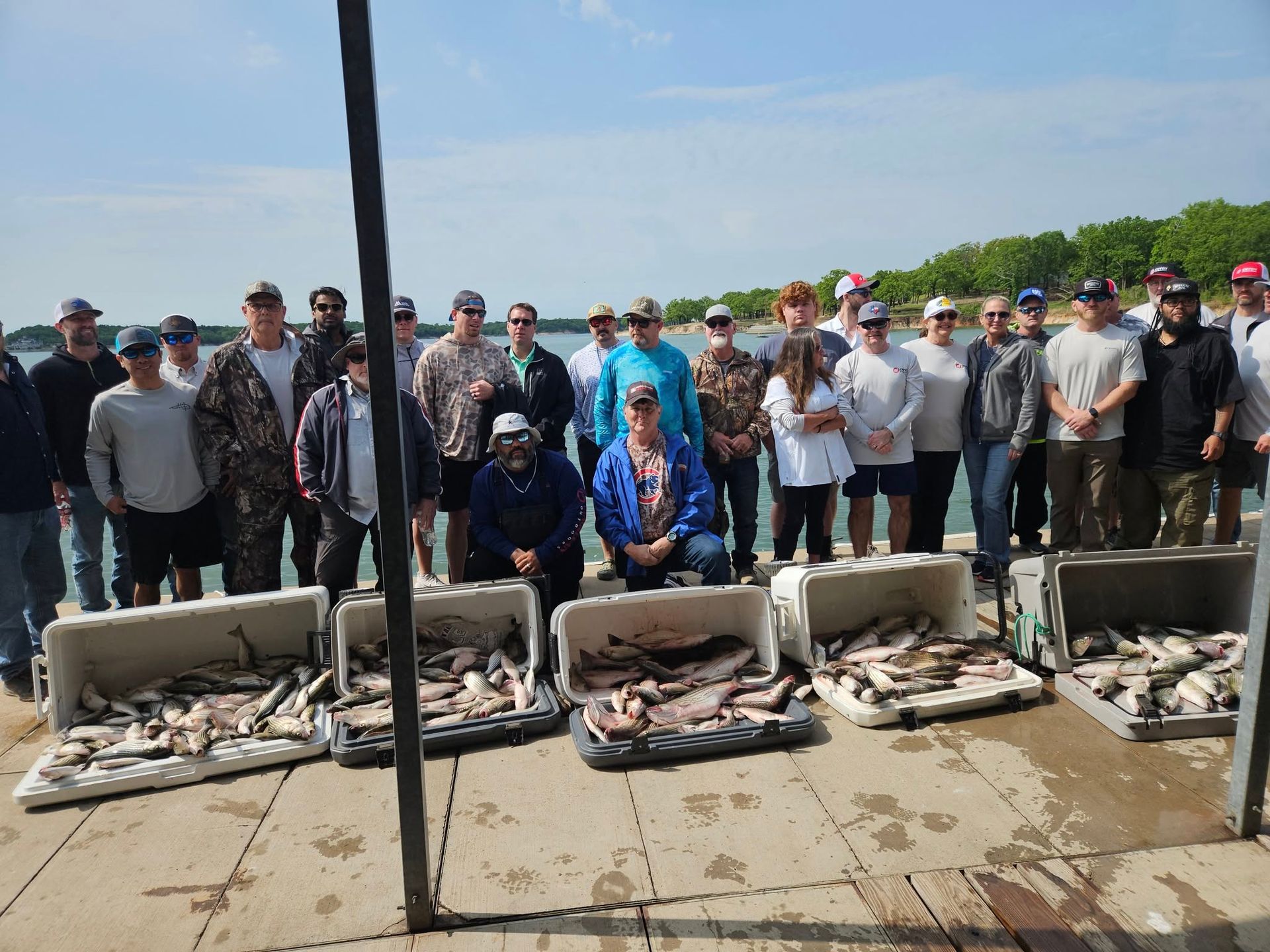 A large group stands on a pier by a lake behind several open coolers filled with a large catch of fresh fish.