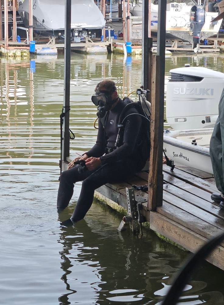 A scuba diver in a black wetsuit sits on a wooden dock at a marina, preparing to enter the water.